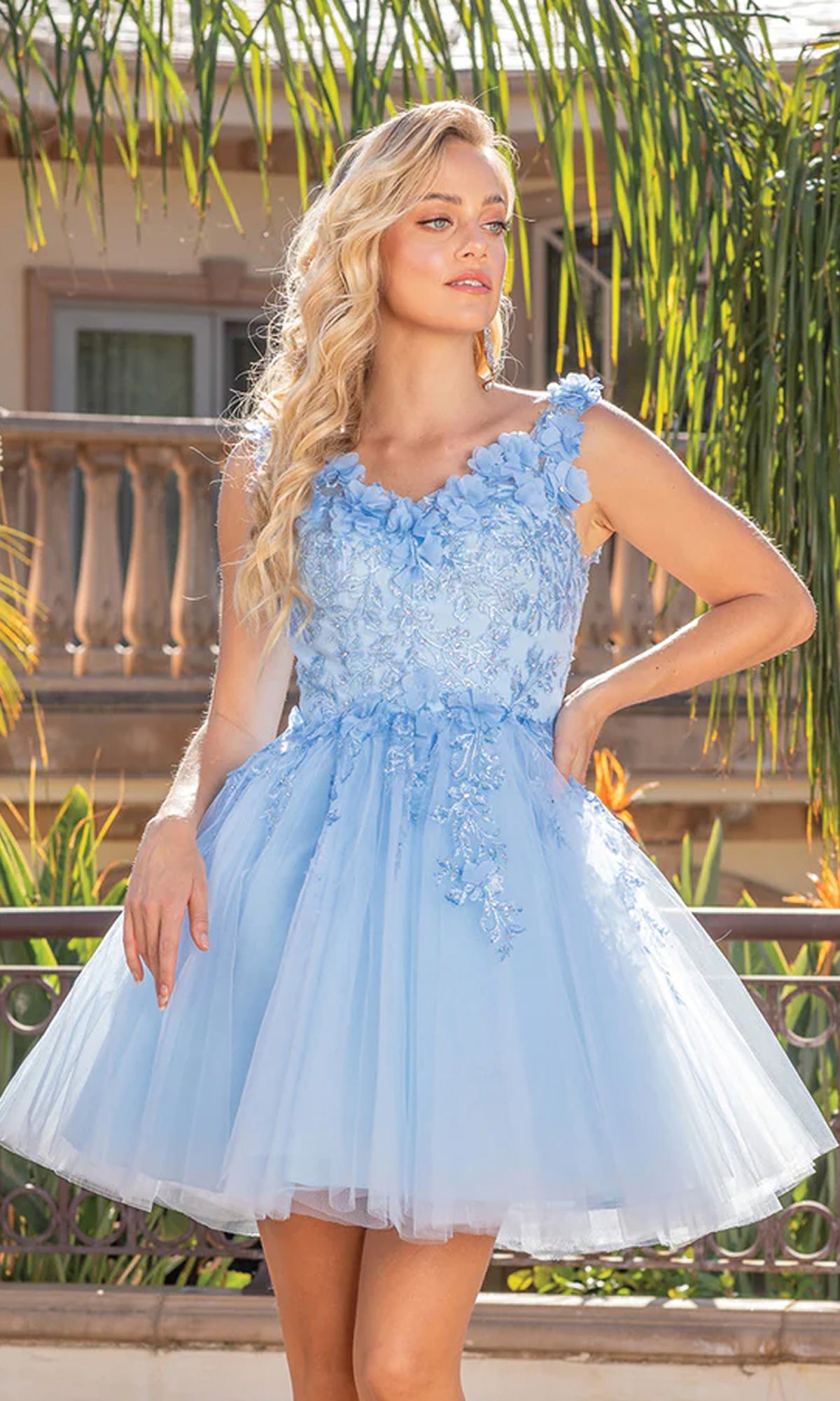 A woman with long blonde hair wears the Corset-Back Short Homecoming Dress with 3D Flowers, a light blue party dress, while standing outdoors in front of palm trees and a balcony.